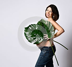 Pregnant girl in jeans, naked to waist. Smiling, covering chest and belly by two green leaves, posing sideways isolated on white