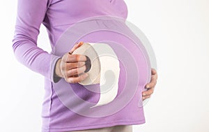 A pregnant girl in a lilac blouse holds toilet paper against the background of her belly. The concept of constipation and diarrhea