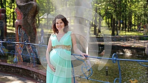 A pregnant girl in a long dress stands in a city Park on a fountain background.