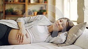 Pregnant girl is lying on bed with closed eyes and stroking her belly with tenderness and love. Healthy pregnancy