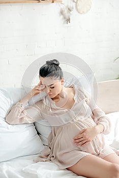 Pregnant girl in nightie having headache and touching belly while sitting on bed