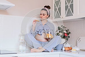 A pregnant girl in pajamas in a good mood drinks tea with cookies in the kitchen