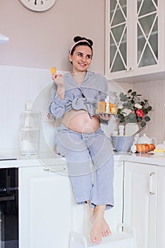 A pregnant girl in pajamas in a good mood drinks tea with cookies in the kitchen