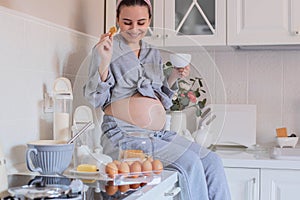 A pregnant girl in pajamas in a good mood drinks tea with cookies in the kitchen