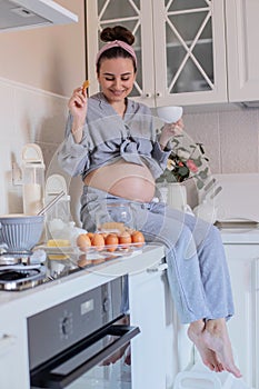 A pregnant girl in pajamas in a good mood drinks tea with cookies in the kitchen