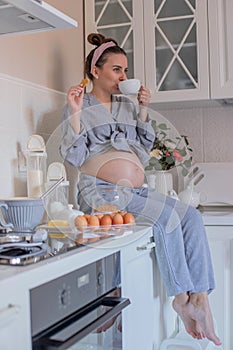 A pregnant girl in pajamas in a good mood drinks tea with cookies in the kitchen