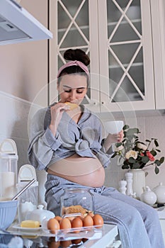 A pregnant girl in pajamas in a good mood drinks tea with cookies in the kitchen