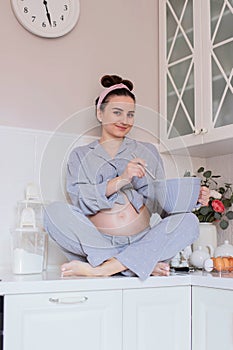 A pregnant girl in pajamas is in a good mood, laughing and cooking sitting on the table in the kitchen