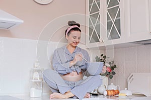 A pregnant girl in pajamas is in a good mood, laughing and cooking sitting on the table in the kitchen