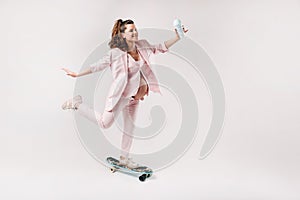 A pregnant girl in a pink suit with a skateboard in her hands stands on a gray background