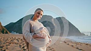Pregnant girl posing seashore with happy smile. Expectant mother standing beach.