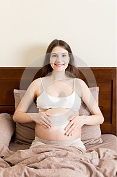 A pregnant girl sits at home on the bed and smears an anti-stretch mark cream on her stomach. Pregnancy, motherhood, preparation