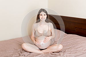 A pregnant girl sits at home on the bed and smears an anti-stretch mark cream on her stomach. Pregnancy, motherhood, preparation