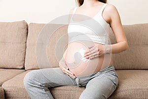 A pregnant girl sits at home on the bed and smears an anti-stretch mark cream on her stomach. Pregnancy, motherhood, preparation