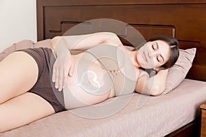 A pregnant girl sits at home on the bed and smears heart an anti-stretch mark cream on her stomach. Pregnancy, motherhood,