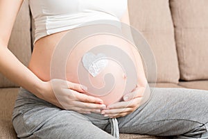 A pregnant girl sits at home on the bed and smears heart an anti-stretch mark cream on her stomach. Pregnancy, motherhood,