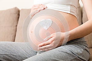 A pregnant girl sits at home on the bed and smears heart an anti-stretch mark cream on her stomach. Pregnancy, motherhood,