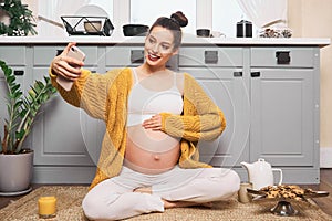 A pregnant girl sits on the kitchen floor and takes a selfie