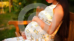 A pregnant girl sits in a park on a bench in a white dress, stroking her belly