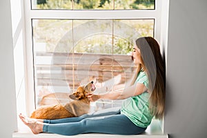 Pregnant girl sitting on the windowsill and hug Corgi dog at home. Young woman play with lovely Welsh Corgi Pembroke in