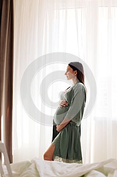 A pregnant girl stands at the window with curtains and looks out the window