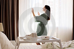 A pregnant girl stands at the window with curtains and looks out the window