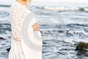 Pregnant girl in a white dress holds her belly in her hands on the background of the sea