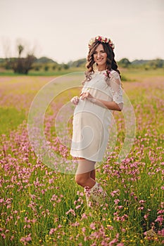 Pregnant happy young woman in floral wreath stand on grass in the outdoor in the field of pink flowers background with. Close up