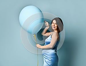 Pregnant lady in blue silk dress or nightie. She is smiling, holding balloon by yellow ribbon, posing sideways on blue background