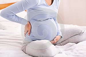 Pregnant Lady Having Backache Sitting On Bed In Bedroom, Cropped
