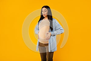 Pregnant lady having painful spasm and touching belly, doing breathing exercises over yellow background