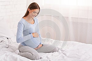 Pregnant Lady Touching Belly Smiling Sitting On Bed At Home