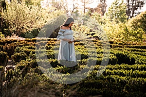 Pregnant Latina woman in a dress standing in lush green sunlit park, hands gently cradling her belly