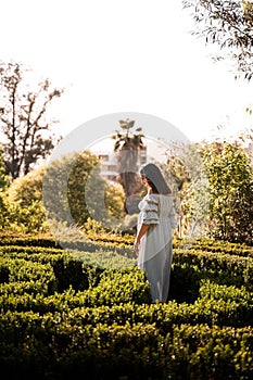 Pregnant Latina woman in a dress standing in lush green sunlit park, hands gently cradling her belly