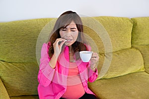 pregnant latina woman taking medicine with headache expression and cup of coffee. On the sofa at home