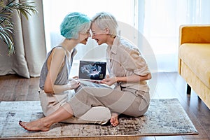 Pregnant lesbian couple with sonogram on floor in living room, side view portrait