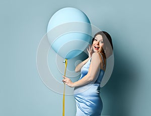 Pregnant model in blue silk dress or nightie. She is smiling, holding balloon by yellow ribbon, posing sideways on blue background