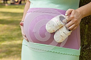 Pregnant mother holding newborn baby's bootie in a park.