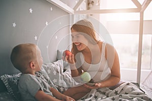 Pregnant mother and little boy son are eating an apple and peach in the bed t home in the morning. Casual lifestyle in