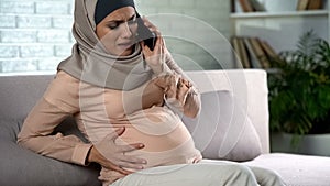 Pregnant muslim female feeling pain, calling emergency smartphone, contractions