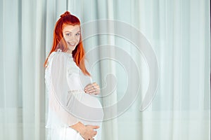 Pregnant. Portrait of young 39 weeks pregnant redhead attractive woman happy smiling standing by the window white curtain dressed