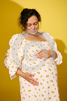 Pregnant pretty woman in sundress, gently caresses her belly, experiencing happy moments of her carefree happy pregnancy
