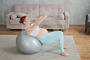 Pregnant red-haired woman doing exercises on fitness ball at home.