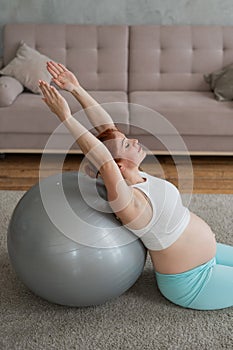 Pregnant red-haired woman doing exercises on fitness ball at home.