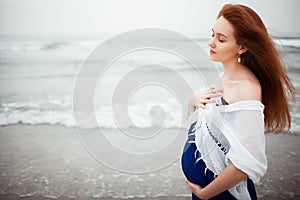 Pregnant redhead girl in a blue dress and a white shawl is on the backdrop of the ocean. Meditating on the sound of the