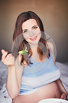 Pregnant smiling girl in T-shirt sit on bed and eat salad with green and red leaves. Young woman keep diet during pregnancy. Healt