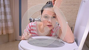 Pregnant tired woman is vomiting in toilet sitting on the floor at home and looking at camera.