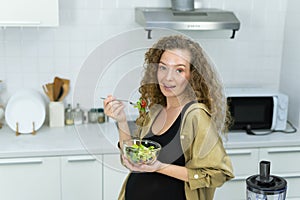 pregnant vegetarian woman smile, happy, stand, eat vegetable, look at camera in kitchen with copy space. beautiful caucasian