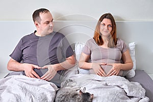 Pregnant wife and overweight husband lie in bed holding their bellies, concept of sympathetic pregnancy