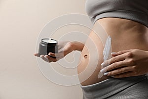 Pregnant woman applying cosmetic product on belly against beige background, closeup